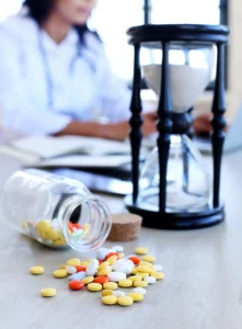 Doctor at his desk writing. On the desk there are pills and a hourglass.