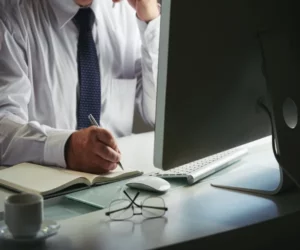 Man writing with a pen in front of a computer the glasses on the desk.